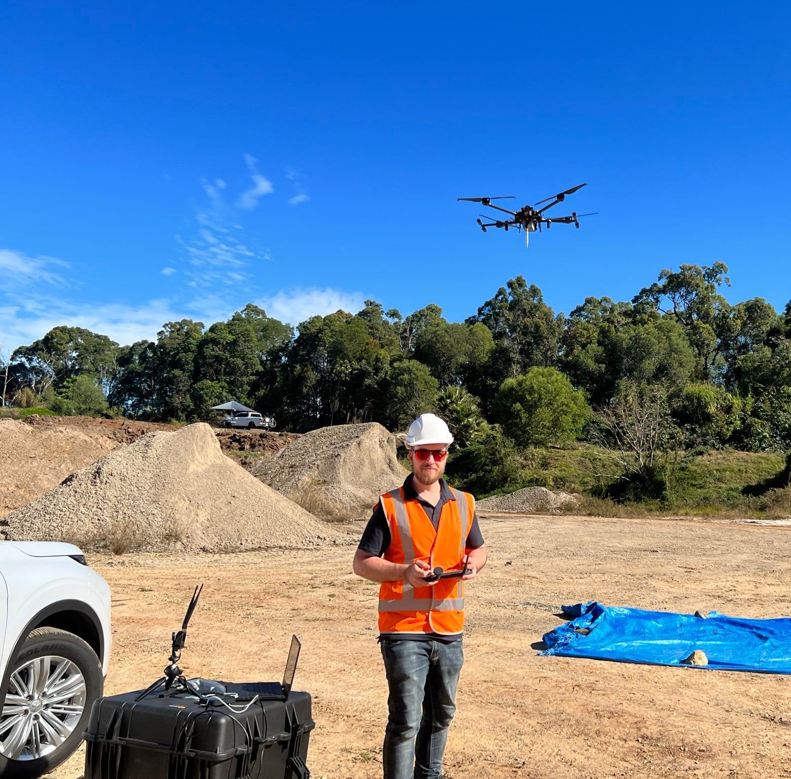 A multirotor drone in flight over a test site, with an operator in high-visibility gear below